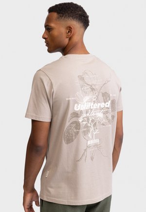 Νεαρός με κοντά μαλλιά που κοιτάζει αριστερά, φοράει μπεζ μπλουζάκι με λευκό λουλουδένιο σχέδιο και το κείμενο "Unfiltered" στην πλάτη, σε λευκό φόντο.