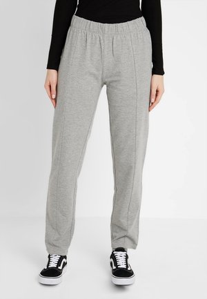 Person iført lysegrå joggingbukser med elastisk talje, sort bluse med lange ærmer og sort-hvide sneakers, stående foran en ensfarvet baggrund.