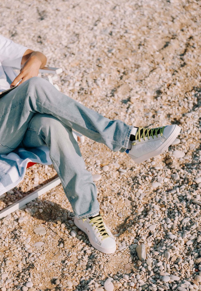 Personne assise sur une chaise de plage rocheuse, jambes croisées, portant un jean bleu clair et des baskets blanches avec des lacets verts.