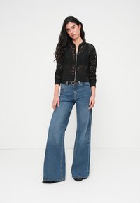 Μαύρο δαντελένιο bomber μπουφάν με μακριά μανίκια και ριμπ τελείωμα, συνδυασμένο με τζιν παντελόνι μπλε χρώματος, ψηλόμεσο και καμπάνα. Απλά μαύρα παπούτσια.