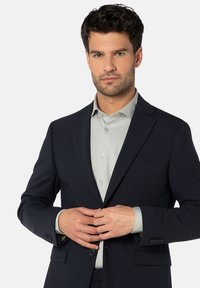 Marineblazer mit Reverskragen, Ein-Knopf-Verschluss und zwei vorderen Taschen. Kombiniert mit einem hellgrauen Hemd mit Kragen und Knöpfen.