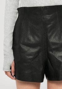 Pantaloni corti neri in pelle a vita alta indossati con un top grigio chiaro a maniche lunghe che mostra un'apertura per il pollice sulla manica e una mano sinistra di lato.