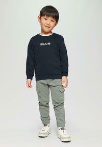 Dunkelblaues Sweatshirt mit dem Text "SLVR", hellgraue Cargo-Hosen mit Taschen und weiße Sneakers mit beigen Akzenten; stehendes Kind mit kurzen Haaren.