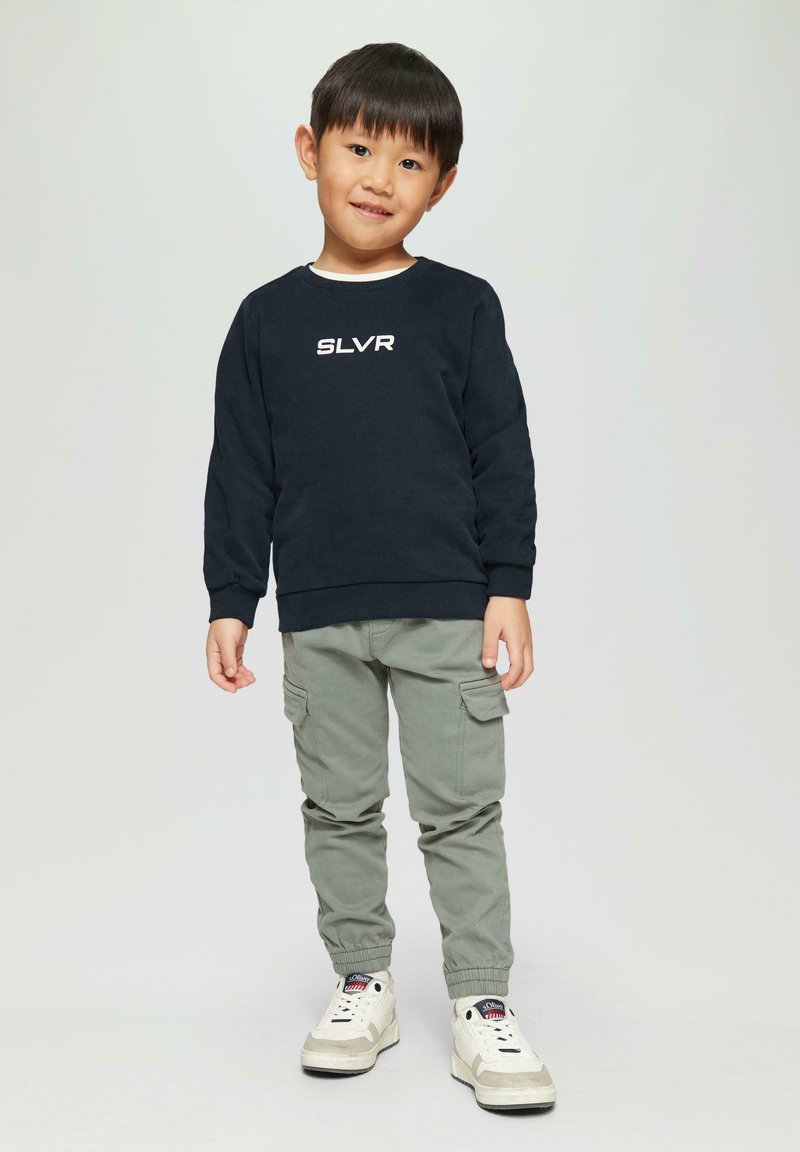 Dunkelblaues Sweatshirt mit dem Text "SLVR", hellgraue Cargo-Hosen mit Taschen und weiße Sneakers mit beigen Akzenten; stehendes Kind mit kurzen Haaren.