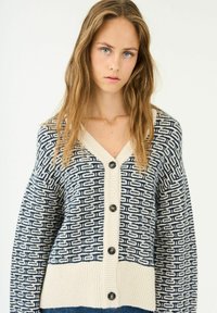 Cardigan a maglia in crema e blu navy con motivo geometrico, scollo a V, maniche lunghe e cinque grandi bottoni scuri sul davanti.