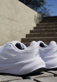 Baskets de sport blanches avec une tige texturée, trois bandes réfléchissantes et une semelle épaisse et striée. Positionnées sur des marches en pierre à l'extérieur.