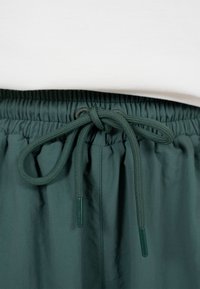 Mørkegrønne bukser med elastik i taljen og bindebånd under en hvid top, der viser stoffets tekstur og syningsdetaljer.