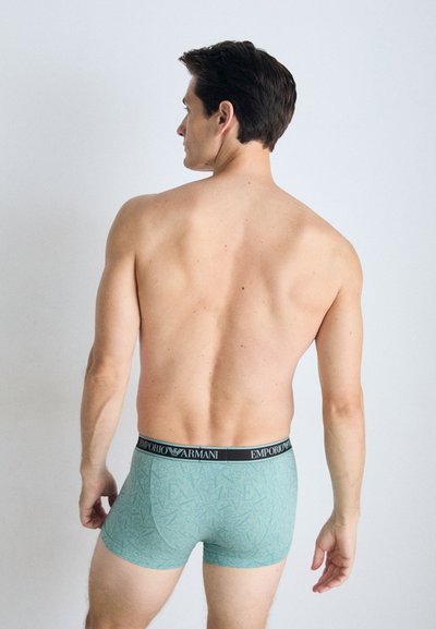 Boxeri scurți cu model verde și talie neagră, pe care se află logo-ul "EMPORIO ARMANI". Țesătura pare netedă și ajustată.