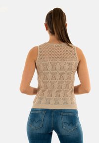 Top de punto sin mangas color beige con un patrón geométrico detallado, escote acanalado y silueta ajustada. Combinado con jeans azules con detalles en los bolsillos.