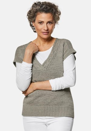 Khakifarbene Strickweste mit V-Ausschnitt, strukturierten Details und kurzen Ärmeln, kombiniert mit einem weißen Langarmshirt und weißen Hosen.