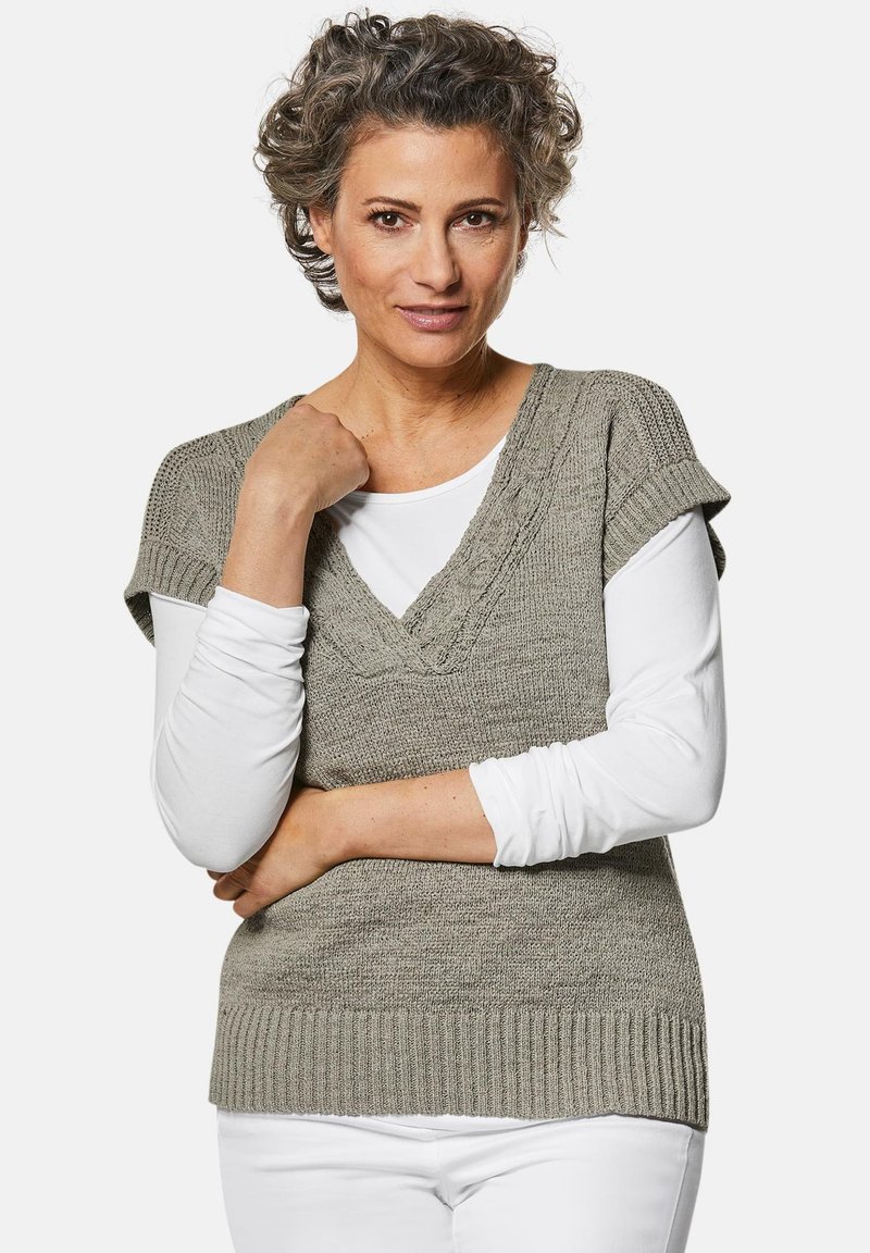 Khakifarbene Strickweste mit V-Ausschnitt, strukturierten Details und kurzen Ärmeln, kombiniert mit einem weißen Langarmshirt und weißen Hosen.