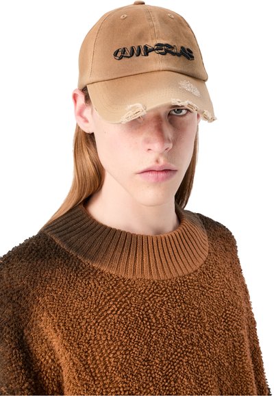 Tan baseball cap with frayed brim and black embroidered "CAMPER" logo. Worn with a textured brown pullover featuring a ribbed neckline.