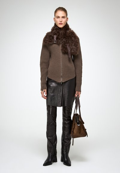 Donna che indossa un maglione marrone a coste con zip e colletto di pelliccia, gonna in pelle nera e pantaloni neri, stivali neri, mentre tiene una borsa in suede marrone.