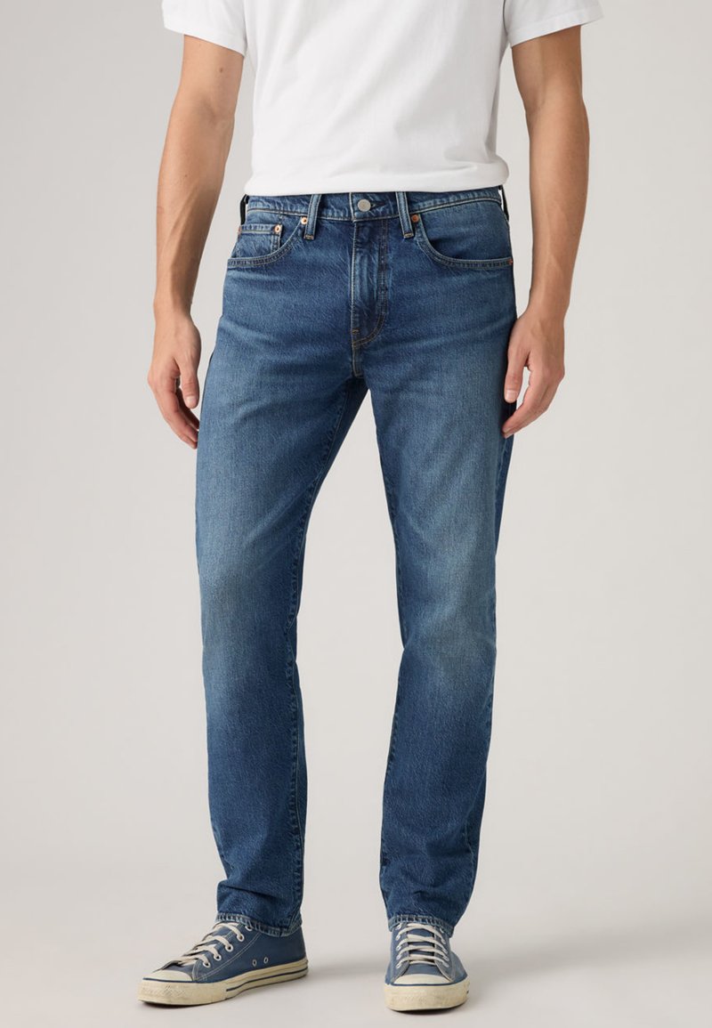 Blue denim jeans with a straight-leg cut, featuring five pockets, metal button closure, and slight fading. Paired with blue sneakers.