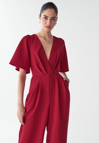 Vrouw met opgestoken haar draagt gouden oorbellen en een rode jumpsuit met een diepe V-hals en brede korte mouwen, staand tegen een witte achtergrond.