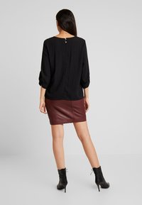 Blusa nera con texture, maniche a tre quarti e dettaglio dei bottoni sul retro. Abbinata a una mini gonna in pelle bordeaux e stivali alla caviglia neri.