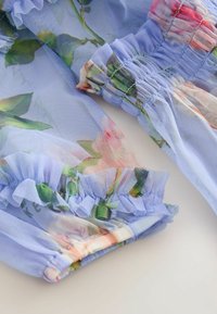 Helles blaues Netzgewebe mit floralem Druck, mit Rüschensäumen und elastischen Bündchen. Die Textur ist zart und durchsichtig.