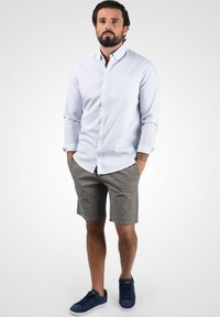 Ljusblå skjorta med knapp-up och bröstficka, parat med grå rutiga shorts. Modellen har på sig marinblå sneakers och står framför en neutral bakgrund.