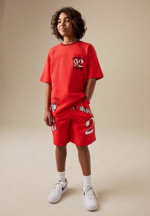 Pois lokkis juustega, kellel on punane suur T-särk ja vastavad püksid, millel on valged ja mustad graafikud, valged Nike tossud, seisab ühe käe taskus.