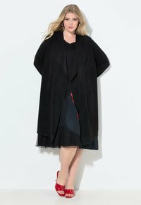 Schwarzer langer Cardigan aus weichem, strukturiertem Stoff mit offenem Vorderteil, geschichtetem Design und weiten Ärmeln, getragen über einem dunklen Kleid und roten High Heels.