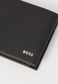 Portefeuille en cuir noir avec une finition texturée, portant le logo argenté "BOSS" à l'avant et des coutures visibles le long des bords.