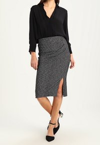 Blouse noire à manches longues associée à une jupe crayon ajustée avec un motif géométrique et une fente sur le côté, portée avec des talons noirs.