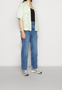 Chemise à manches courtes tie-dye vert clair, débardeur noir, jean bleu taille haute, et baskets de sport argentées avec des accents réfléchissants.