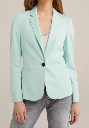 Femme portant un blazer bleu clair sur mesure avec un seul bouton noir, un haut bordé de dentelle et un jean gris sur fond beige.