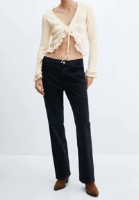 Kremowy, koronkowy, krótkie kardigan z wiązaniem i falbaną na dole, w połączeniu z czarnymi, prostymi dżinsami oraz brązowymi botkami na obcasie.