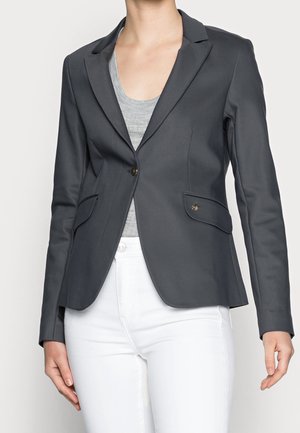 Kvinde iført en tætsiddende, mørkegrå blazer med enkelt knap over en lysegrå top og hvide bukser, stående med armene afslappet.