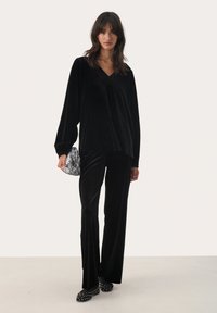 Black velvet top with V-neck and long sleeves paired with matching velvet trousers. Accented with a metallic-patterned clutch and studded flats.