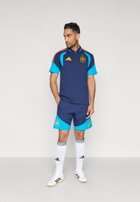 Σετ ποδοσφαίρου σε ναυτικό μπλε και τιρκουάζ, που περιλαμβάνει κοντομάνικο μπλουζάκι με φερμουάρ στο λαιμό, αντίστοιχα σορτς και λευκές κάλτσες με μαύρες ρίγες. Διακριτικά της Adidas εμφανή.