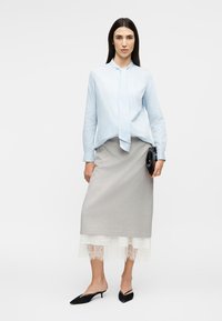 Γυναίκα που φορά μπλούζα με ανοιχτό μπλε ρίγες, γκρι μίντι φούστα με λευκή δαντέλα στο τελείωμα, μαύρα μυτερά τακούνια με λουράκι και κρατάει μαύρο τσαντάκι χειρός.