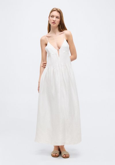 Žena v bielej bezrukávovej maxi šaty s tenkými ramienkami a hlbokým výstrihom do V, obutá v béžových tkaných sandáloch, stojaca na jednoduchom pozadí.