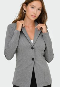 Femme ajustant le col d'un blazer gris cintré avec deux boutons noirs, portant des boucles d'oreilles en or en forme de créole et un haut blanc en dessous.
