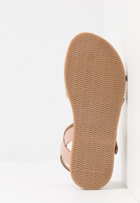 Sanda ligera de color marrón claro con una suela de goma texturizada, que cuenta con correas elásticas sobre el pie. La suela tiene un patrón ondulado para mayor tracción.