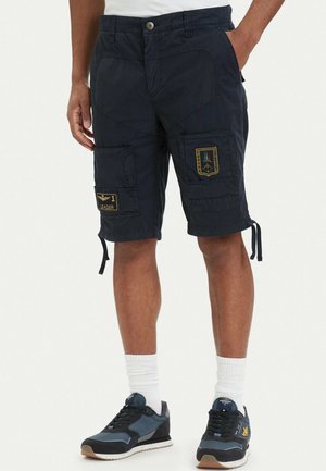 Uomo che indossa pantaloncini cargo scuri con toppe, calzini bianchi e scarpe da ginnastica nero-blu, in piedi con le mani in tasca davanti a uno sfondo semplice.