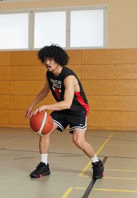 Basketbola spēlētājs driblē bumbu melnā bez piedurknēm kreklā ar sarkaniem akcentiem, melnos šortos un melnās kedās ar sarkaniem elementiem sporta zāles grīdā.