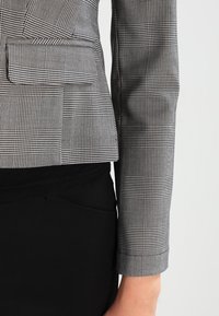 Šedý károvaný blazer se strukturovanými rameny, jedním prsním kapsou, hladkým materiálem a černými akcenty. Detailní záběr na design a vzor rukávu.