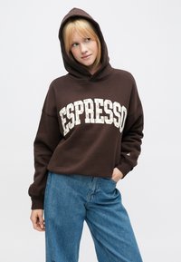 Sudadera con capucha marrón con un gran texto "ESPRESSO" en blanco, delineado por un patrón floral. Combinada con vaqueros de pierna ancha de color azul claro.