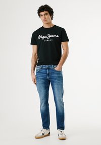 Crna kratka majica s kratkim rukavima sa bijelim logotipom "Pepe Jeans London", u kombinaciji s plavim trapericama. Bijele tenisice s višebojnim detaljima.