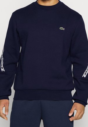 Mann trägt ein marineblaues Lacoste-Sweatshirt mit grünem Krokodil-Logo auf der Brust und weißem Textdesign auf den Ärmeln.