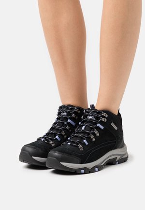 Jambes portant des bottes de randonnée noires avec des détails gris et bleus, lacées, debout sur un fond blanc.
