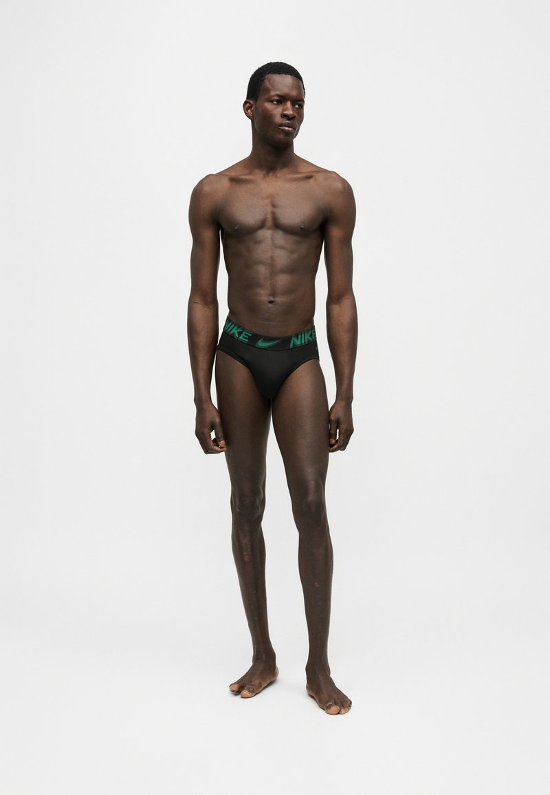 Αθλητικός άνδρας με σκούρο δέρμα, όρθιος ξυπόλητος, φοράει μαύρα εσώρουχα Nike με πράσινη λωρίδα μέσης με λογότυπο σε απλό φωτεινό φόντο.