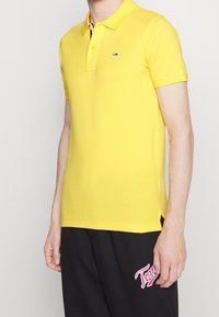 Gul polo skjorta med krage, korta ärmar och en liten logotyp på bröstet. Tyget verkar slätt med en klassisk passform.