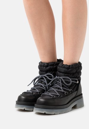 Bottes d'hiver noires rembourrées avec semelles épaisses en caoutchouc gris et lacets noirs à rayures blanches portées sur des jambes nues sur fond blanc.