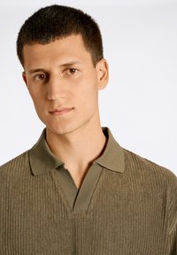 Polo shirt a coste verde oliva con colletto e scollatura a V. La texture è morbida con linee verticali visibili. Nessun motivo o accentuazione.