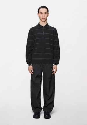 Muž stojí oblečený v čiernom tričku s dlhým rukávom s tenkými vodorovnými pruhmi, voľných čiernych nohaviciach a čiernych topánkach na bielom jednoliatom pozadí.