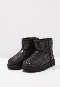 Černé kotníkové boty vyrobené z texturovaného materiálu s lesklým tečkovaným vzorem, s měkkou vnitřní podšívkou a plochou černou podrážkou.