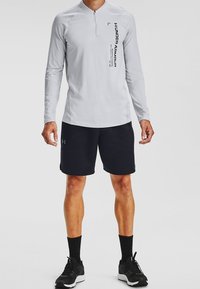 Mies, joka seisoo päällään vaaleanharmaa pitkähihainen Under Armour -vartalo-zip-takki, jossa on pystysuora logo, mustat shortsit, mustat sukat ja mustat urheilukengät.